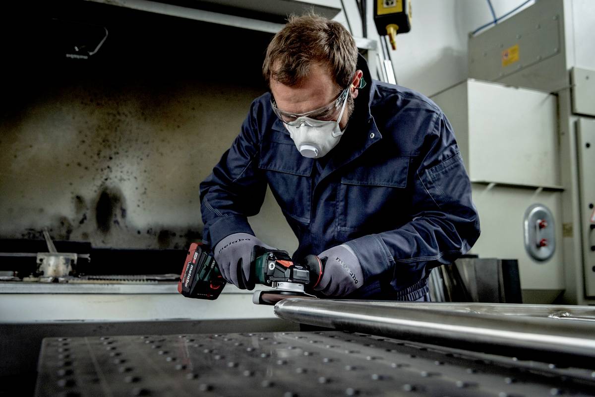 Ein Mann arbeitet mit Schutzbrille und Maske in einer Werkstatt an einer Metallstange mit einem Akkuschrauber.
