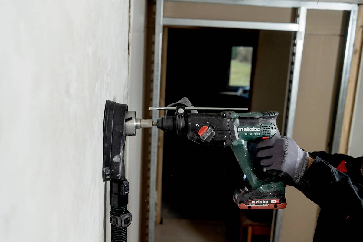 Eine Bohrmaschine durchbricht eine weiße Wand, gehalten von einer Person mit Arbeitshandschuhen. Ein Gebäude im Bauhintergrund.