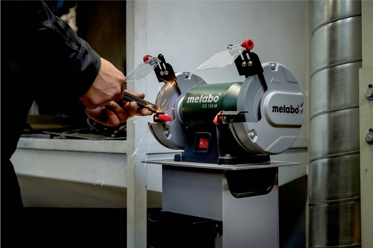 Eine Person schleift ein Werkzeug an einem Metabo Tischschleifer mit Funkenbildung im Hintergrund, in einer Werkstattumgebung.