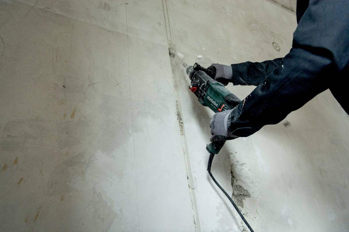 Eine Person trägt Schutzhandschuhe und benutzt eine elektrische Bohrmaschine, um eine vertikale Linie in eine Betonwand zu bohren.