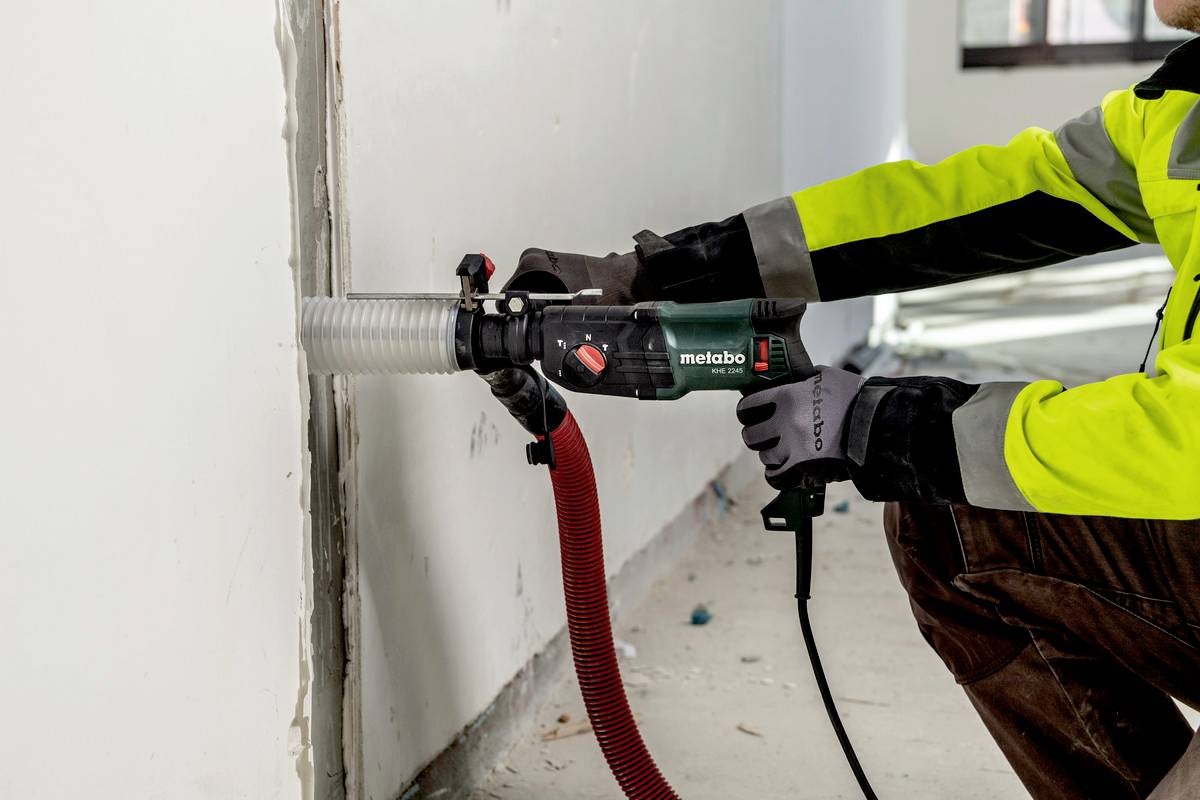 Eine Person in Arbeitskleidung benutzt ein elektrisches Werkzeug mit einem Saugschlauch, um in einer Wand zu sägen.