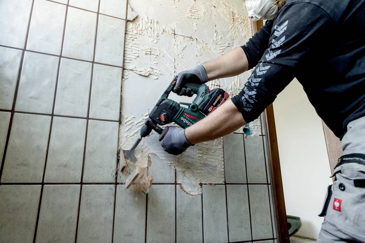 Person entfernt Fliesen von Wand mit Elektrowerkzeug. Sie trägt Schutzkleidung und Maske. Die entfernten Fliesen zeigen freiliegendes Mauerwerk.