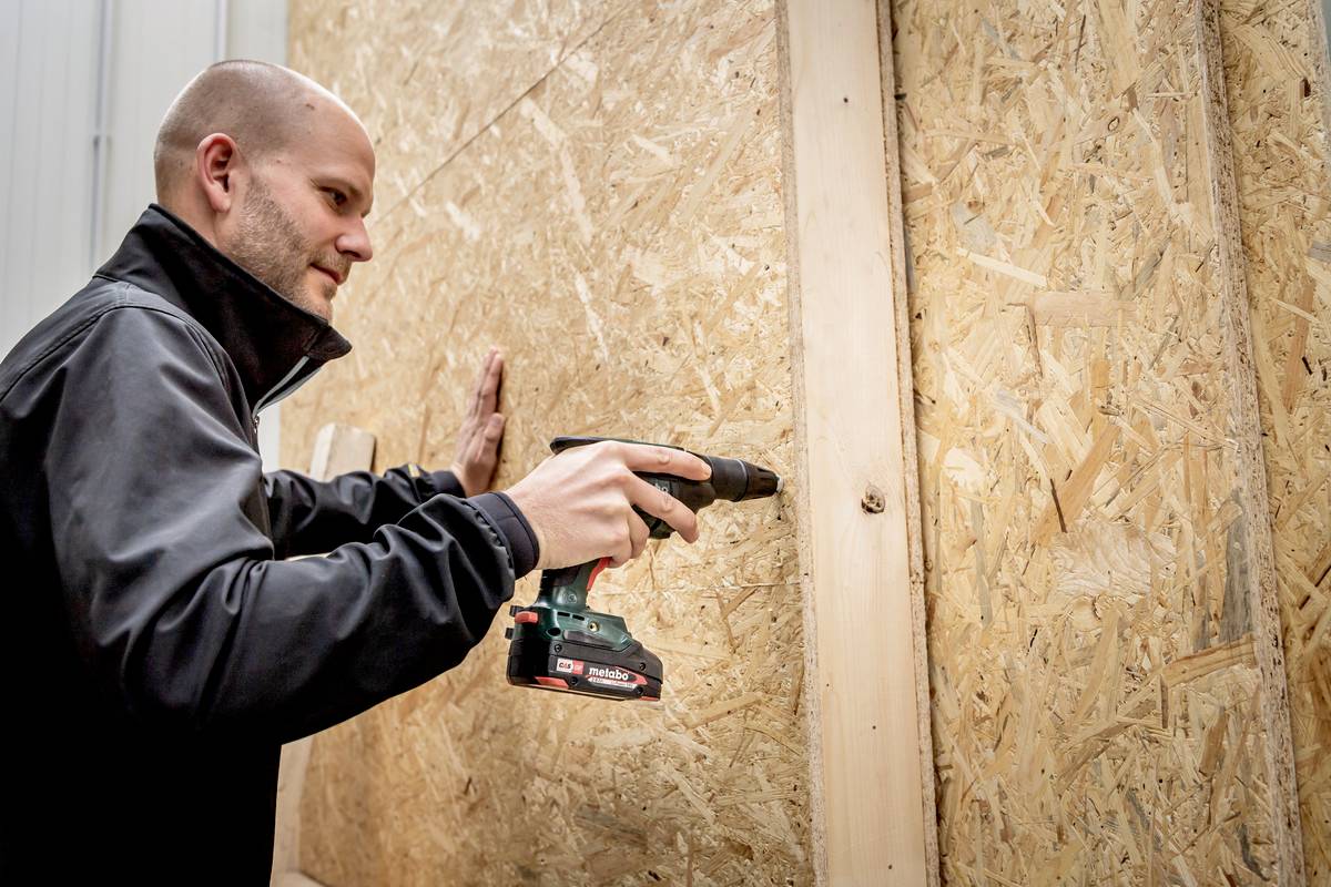 Eine Person befestigt mit einem Akku-Bohrschrauber eine Holzplatte an einer Wand. Sie trägt eine Jacke und arbeitet konzentriert.