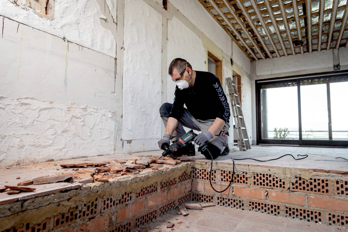 Ein Mann arbeitet mit einem elektrischen Werkzeug auf einer Baustelle. Er trägt eine Schutzmaske. Im Hintergrund sind Leitern und Fenster.