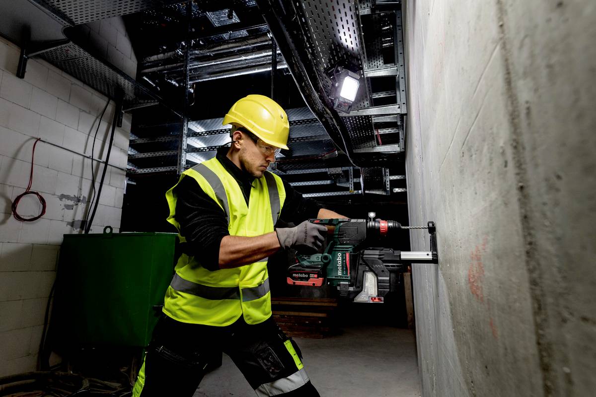 Ein Bauarbeiter in Schutzkleidung bohrt mit einer elektrischen Bohrmaschine in eine Wand. Umgebung ist eine Baustelle mit Rohstruktur.