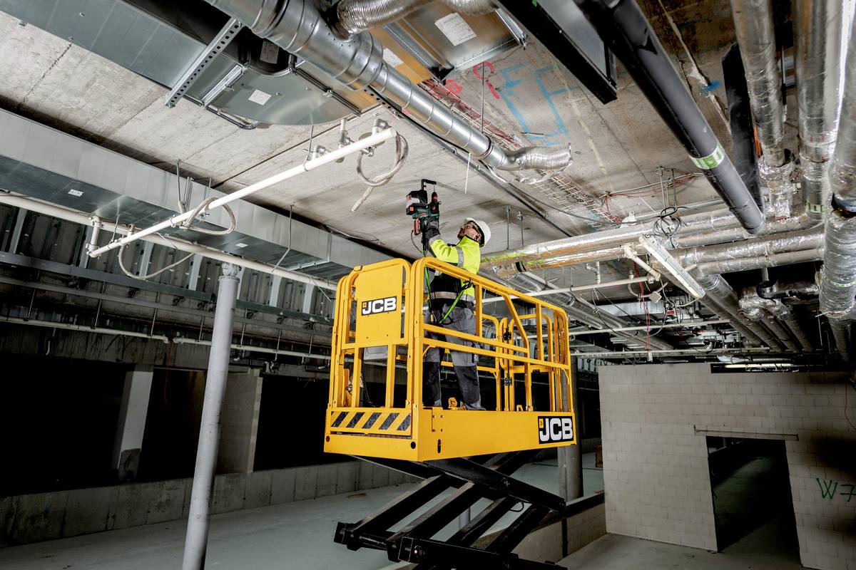 Ein Arbeiter im gelben Kran hebt sich zur Decke eines Gebäudes, um an Kabeln und Rohrleitungen zu arbeiten. Sicherheitsausrüstung wird getragen.