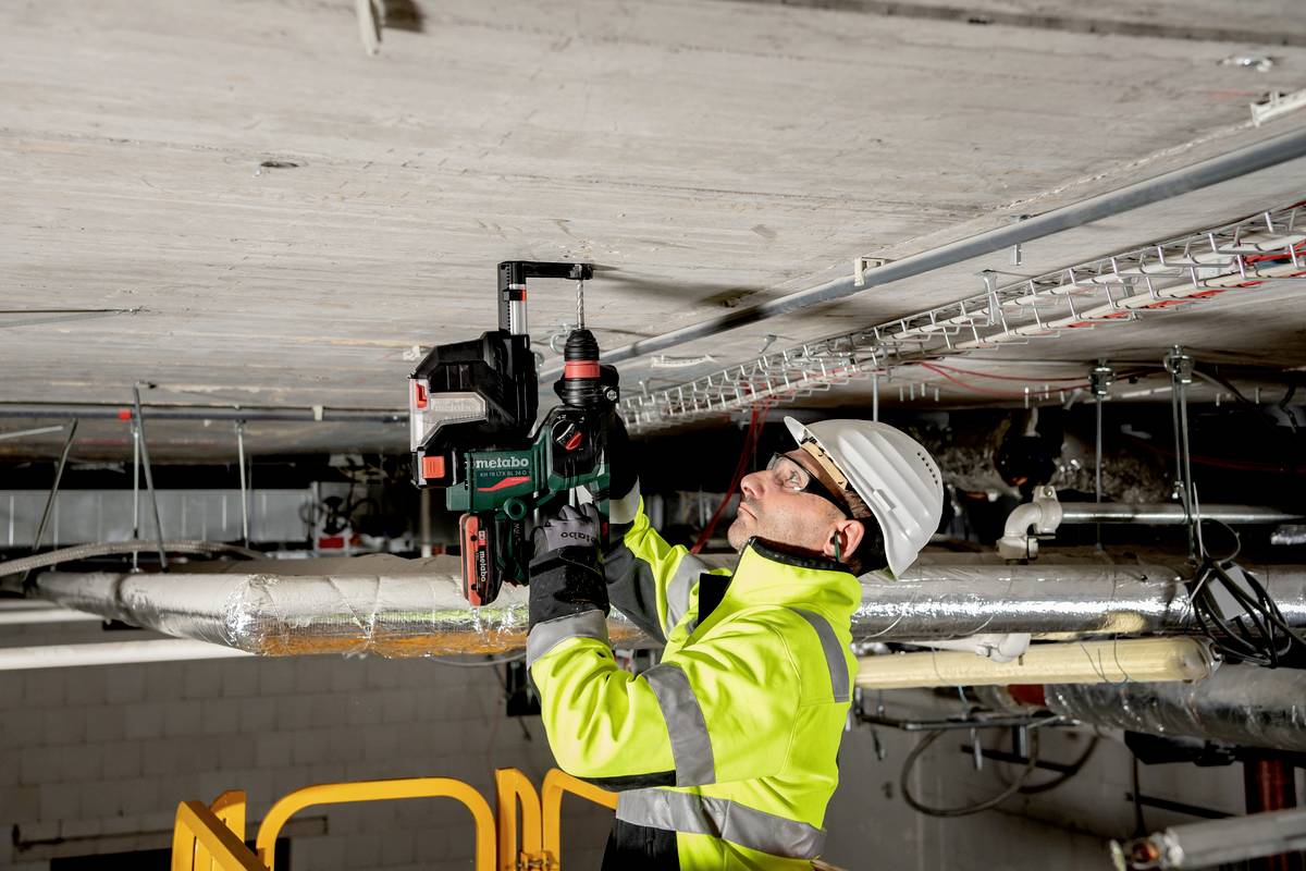 Arbeiter im Bauhelm und gelber Jacke befestigt mit einer Bohrmaschine etwas an der Decke. Im Hintergrund sind Rohre und Kabel sichtbar.
