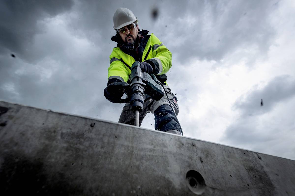 Arbeiter in Schutzkleidung benutzt eine Elektrowerkzeugbohrmaschine auf einer Baustelle, vor bewölktem Himmel.