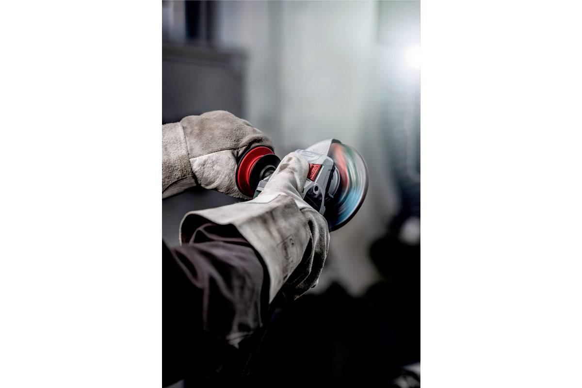 Eine Person schleift mit einer Schleifmaschine Metall. Sie trägt Schutzkleidung und Handschuhe, arbeitet in einer Werkstattumgebung.