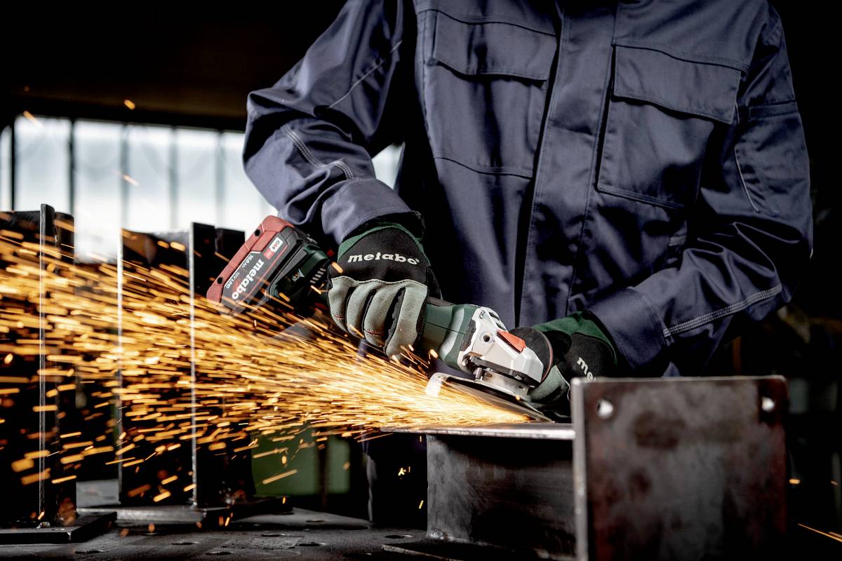 Eine Person schleift Metall mit einer elektrischen Schleifmaschine. Funken springen von der Metalloberfläche ab. Die Person trägt Schutzkleidung.