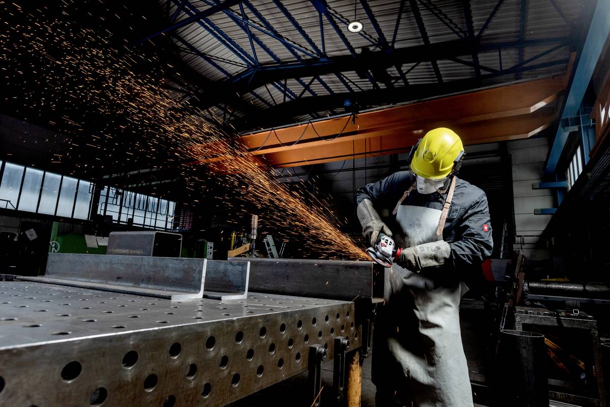 Ein Arbeiter in Schutzkleidung schleift Metall mit einem Winkelschleifer in einer Fabrikhalle, Funken fliegen zur Seite.