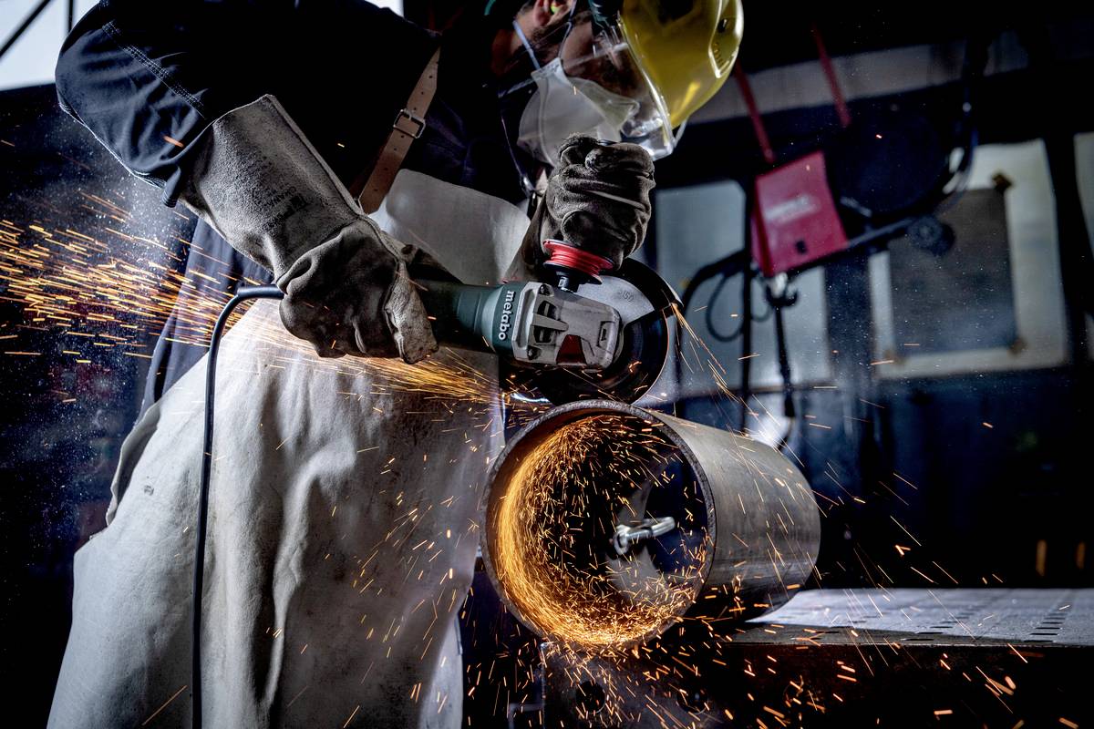 Ein Arbeiter mit Schutzhelm und Schürze schleift einen Metallzylinder mit einer Winkelschleifmaschine, Funken sprühen.
