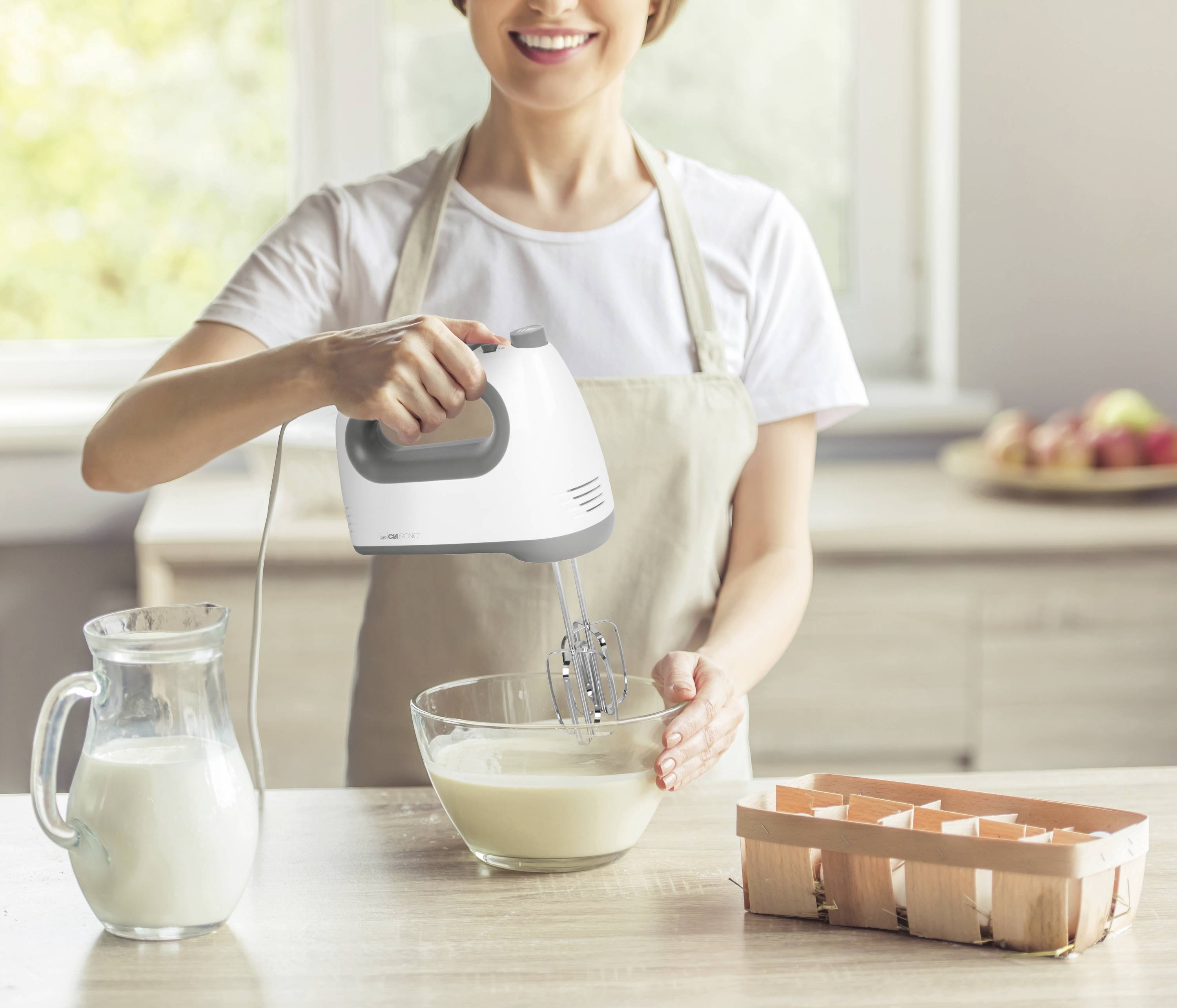 Frau rührt mit einem Handmixer Teig in einer Schüssel in einer Küche. Auf dem Tisch stehen eine Milchkanne und eine Eierkartonschachtel.