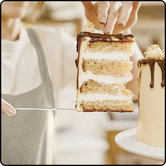 Eine Person schneidet ein Stück mehrschichtige Torte mit Schokoladenglasur ab. Das Umfeld ist eine helle, gemütliche Küche.