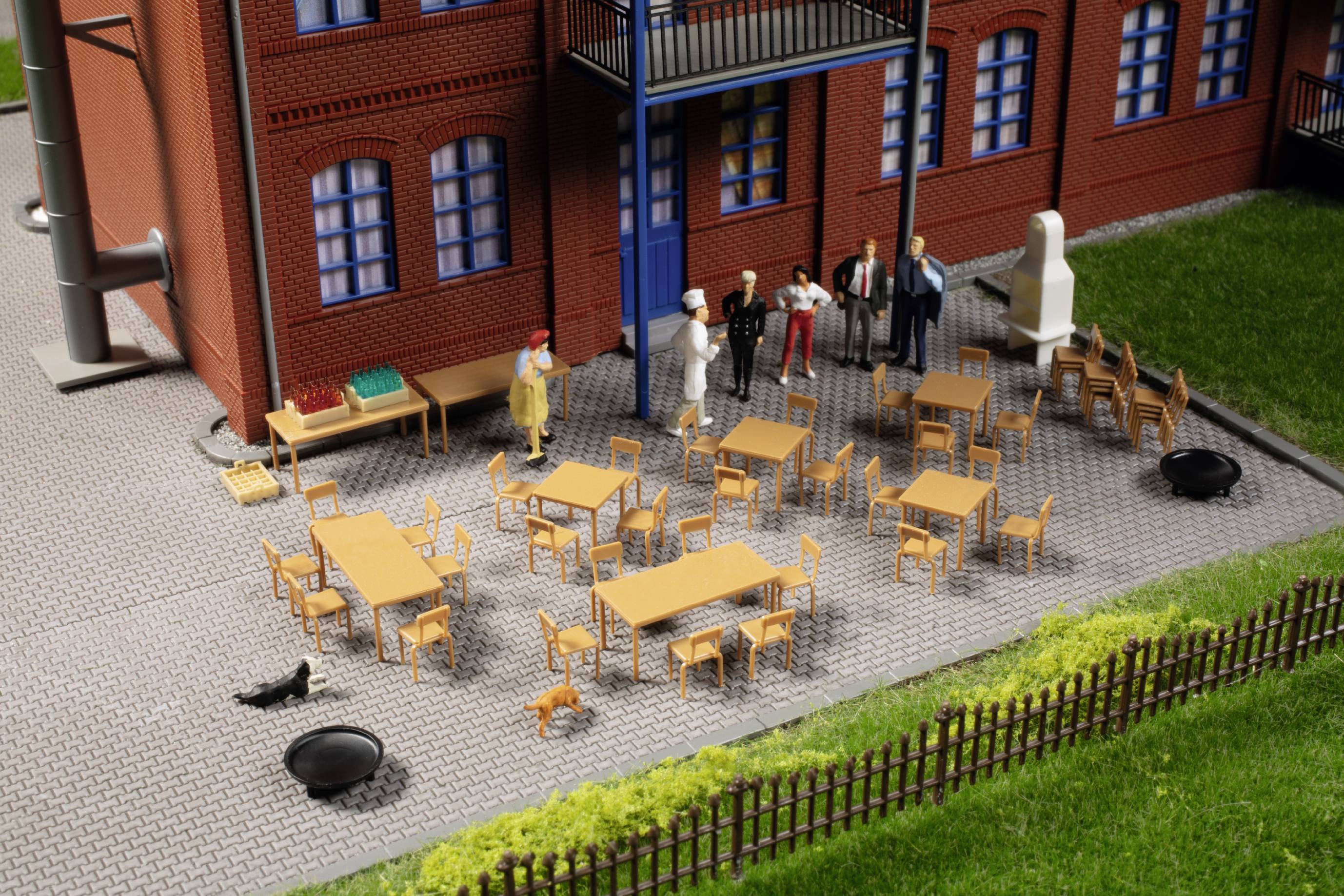 Menschenfiguren stehen auf einer Terrasse mit mehreren Tischen und Stühlen vor einem Backsteingebäude. Ein Rabe und eine Katze sind in der Nähe.