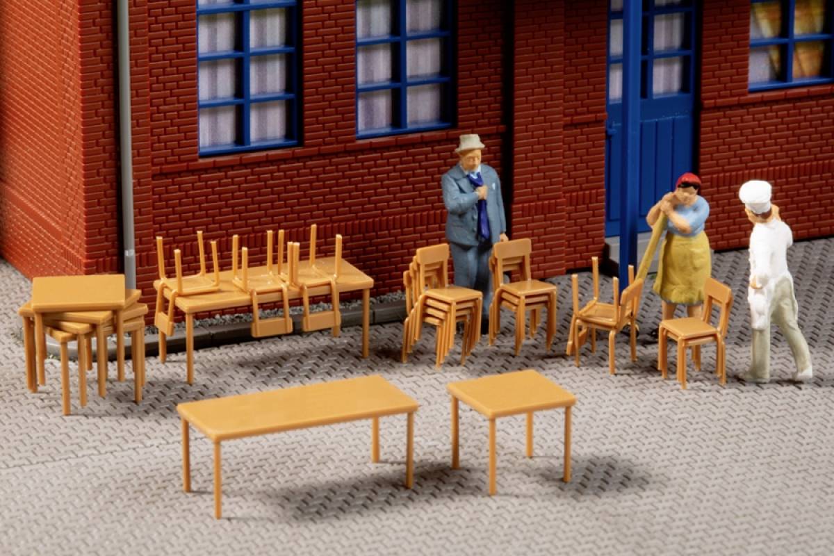 Miniaturmodell zeigt drei Figuren in Diskussion vor einem Gebäude. Daneben sind gestapelte Stühle und ein Tisch arrangiert.