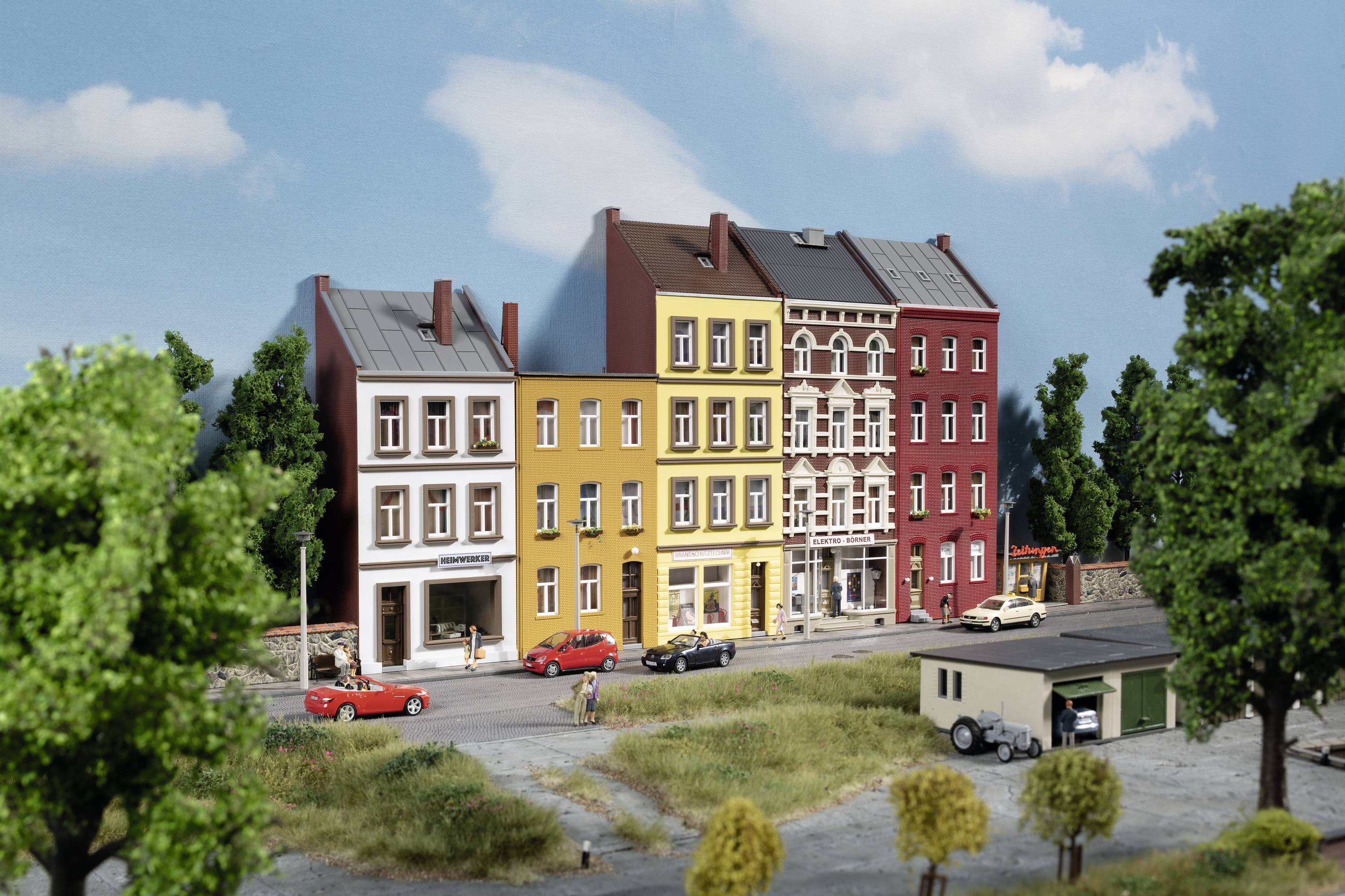 Stadtszene mit drei nebeneinander liegenden, unterschiedlich farbigen Altbauten, mehreren Autos auf der Straße und Bäumen im Vordergrund.