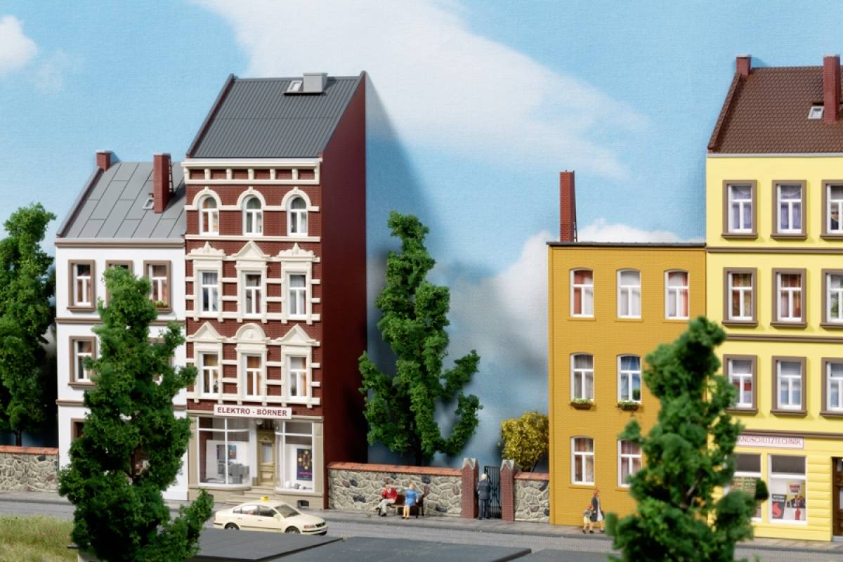 Drei bunte Reihenhäuser mit Geschäften im Erdgeschoss, flankiert von Bäumen, vor einem Hintergrund aus blauem Himmel und Wolken.