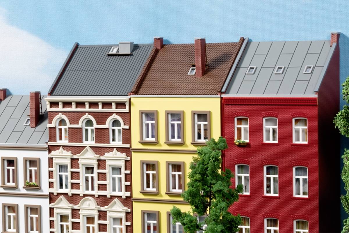 Mehrfarbige Stadthäuser in einer Reihe, jedes mit einzigartiger Fassade und Dach. Ein Baum steht im Vordergrund. Klarer Himmel.