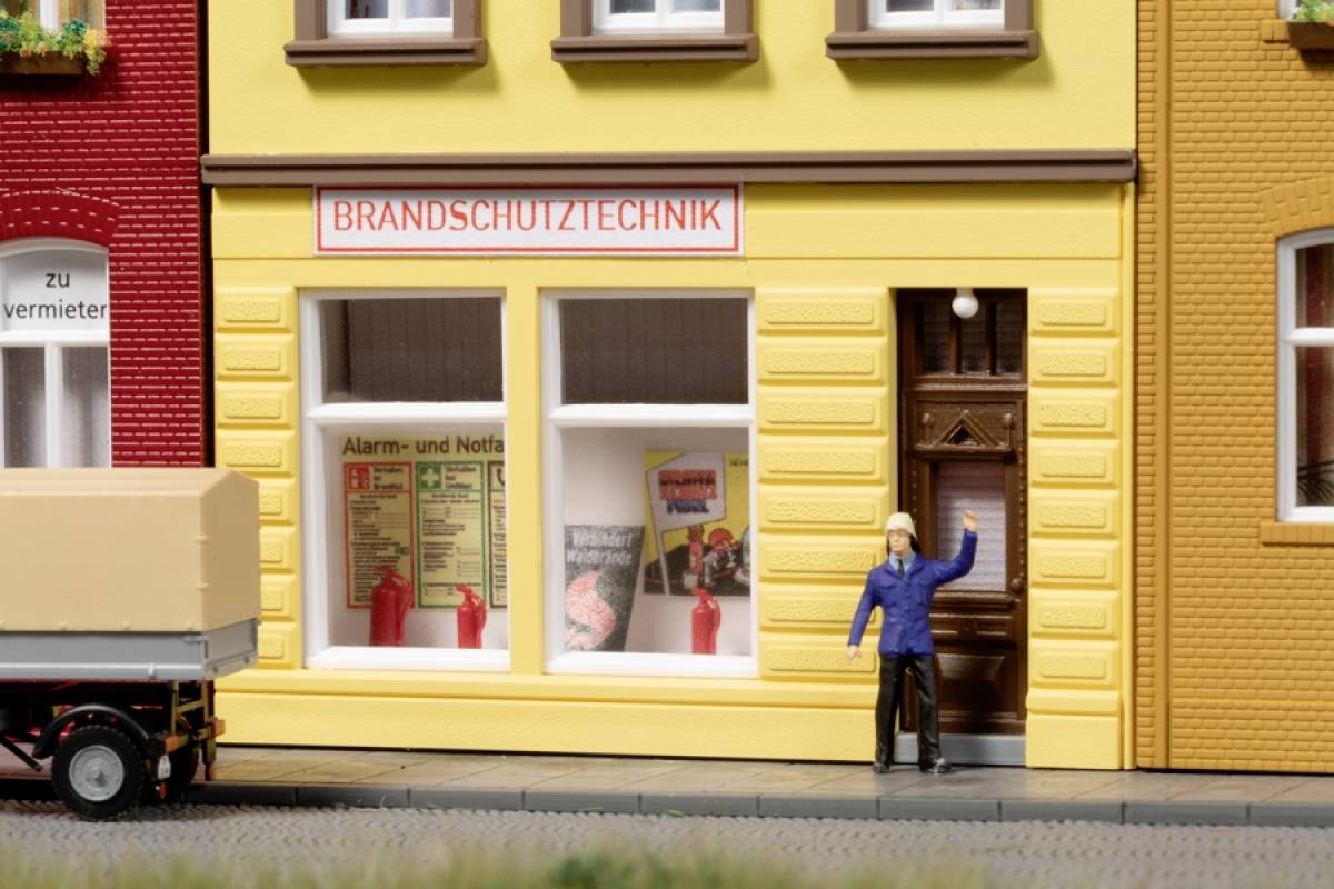 'Brandschutztechnik'-Geschäft mit einem Modell-Lkw und einer Figur im blauen Parka, die zur Tür des gelben Gebäudes geht.