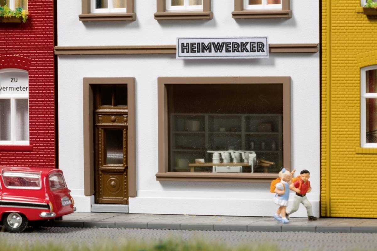 Ein Miniaturmodell eines Straßenzuges mit einem Geschäft namens 'Heimwerker'. Vor dem Laden spielen zwei kleine Figuren. Ein rotes Auto steht links.