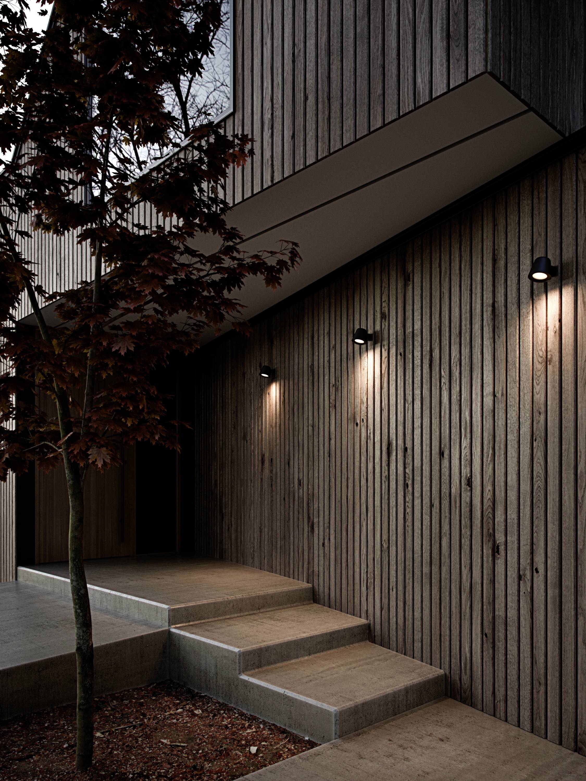 Moderne Gebäudefassade aus Holz mit Beleuchtung. Eine schmale Treppe führt zum Eingang. Daneben steht ein Baum. Dämmerungsstimmung.