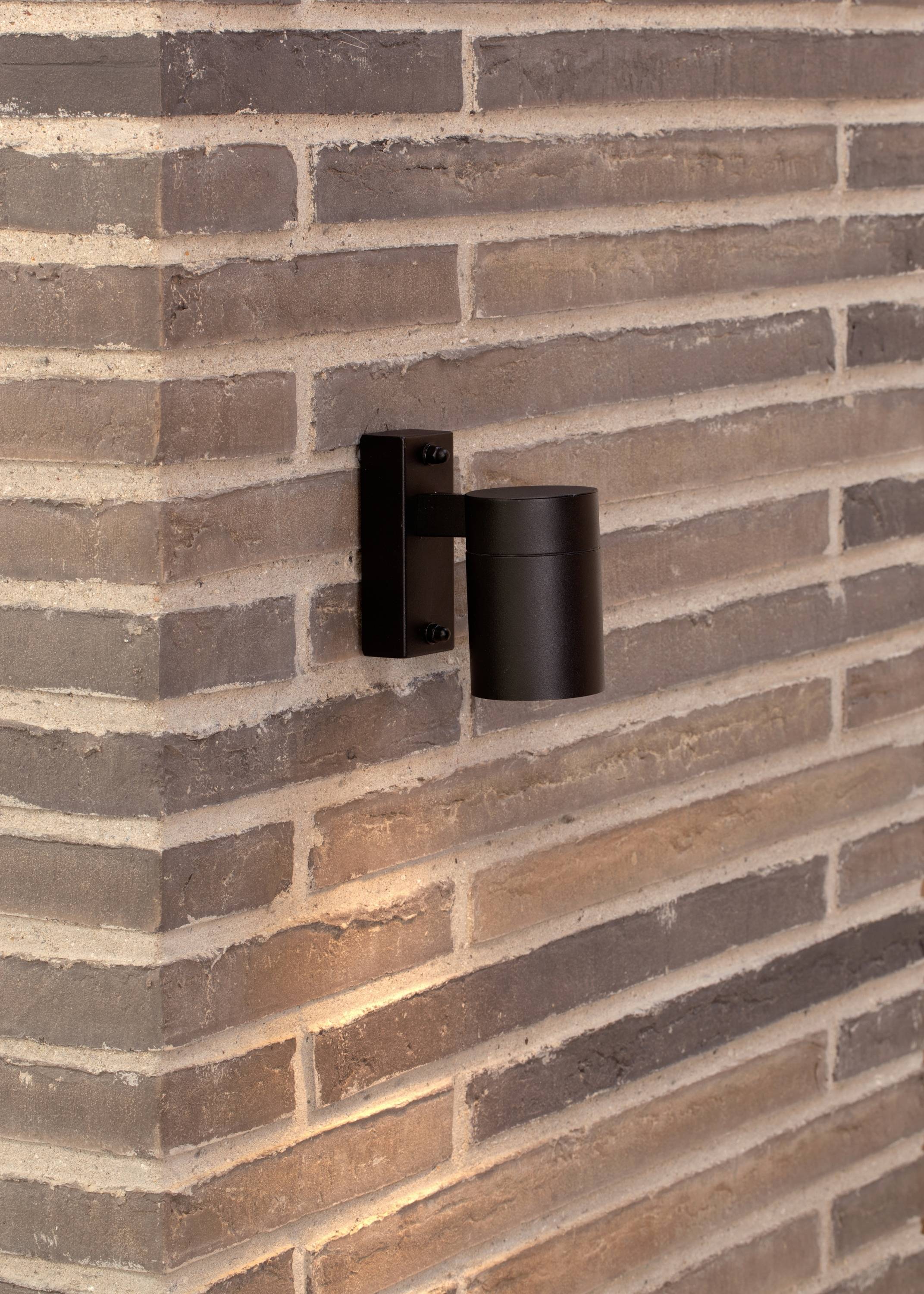 Schwarze Außenleuchte an einer Eckwand aus Ziegelsteinen, beleuchtet den Bereich darunter.