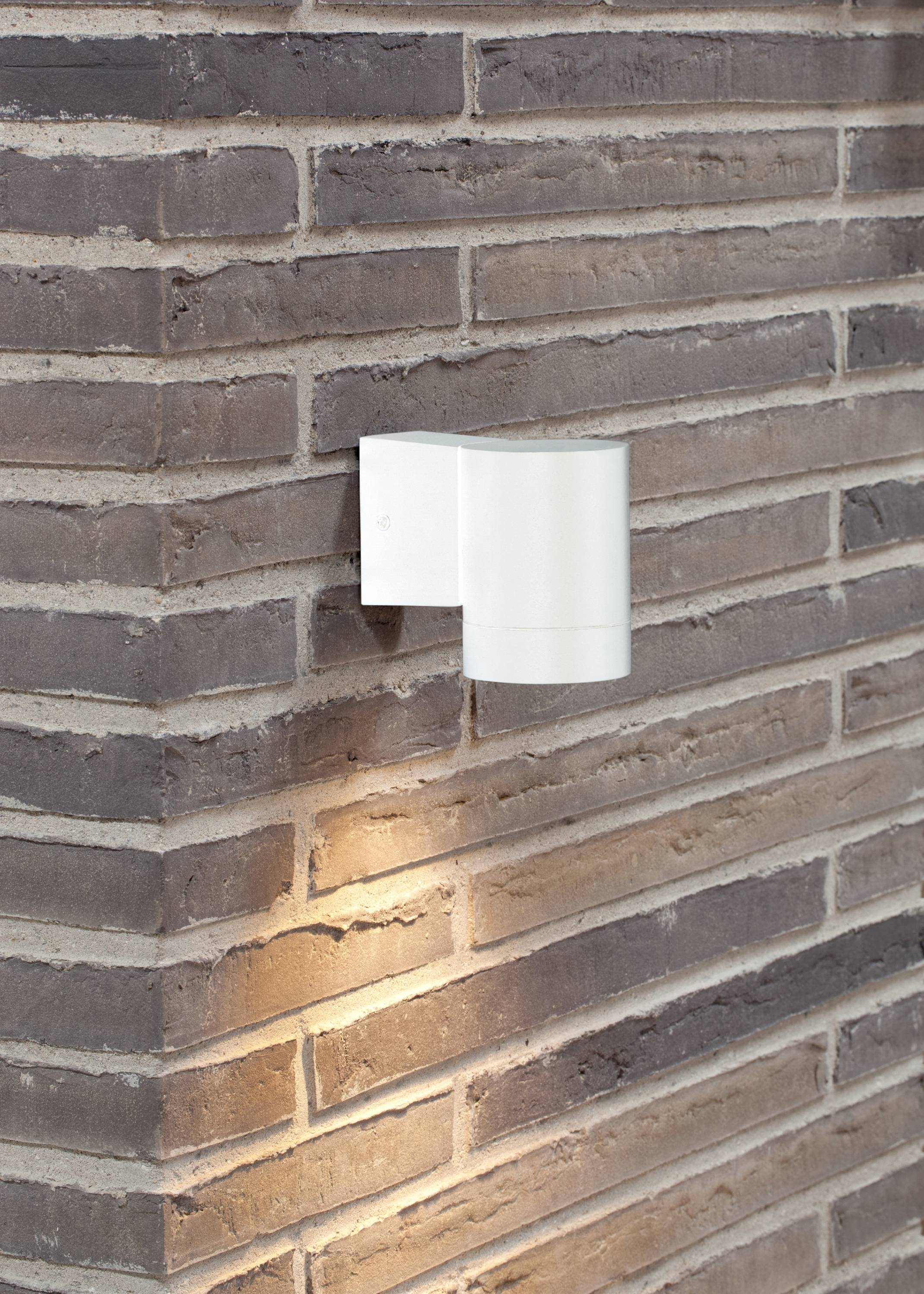 Weiße Wandleuchte an einer Ziegelsteinwand, die nach unten leuchtet und die Wand mit warmem Licht erhellt.