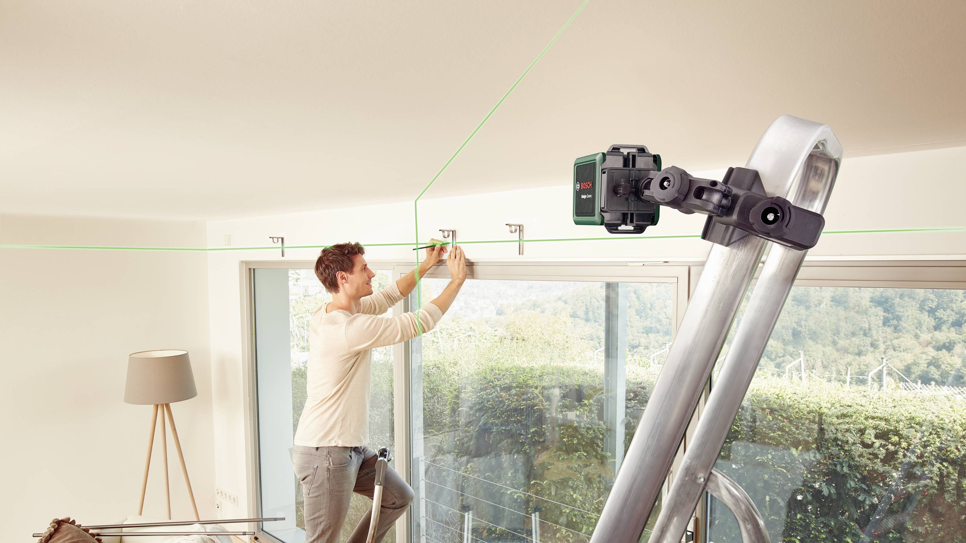 Ein Mann benutzt eine grüne Laser-Wasserwaage, die auf einer Leiter befestigt ist, um Vorhänge über einem großen Fenster zu montieren.