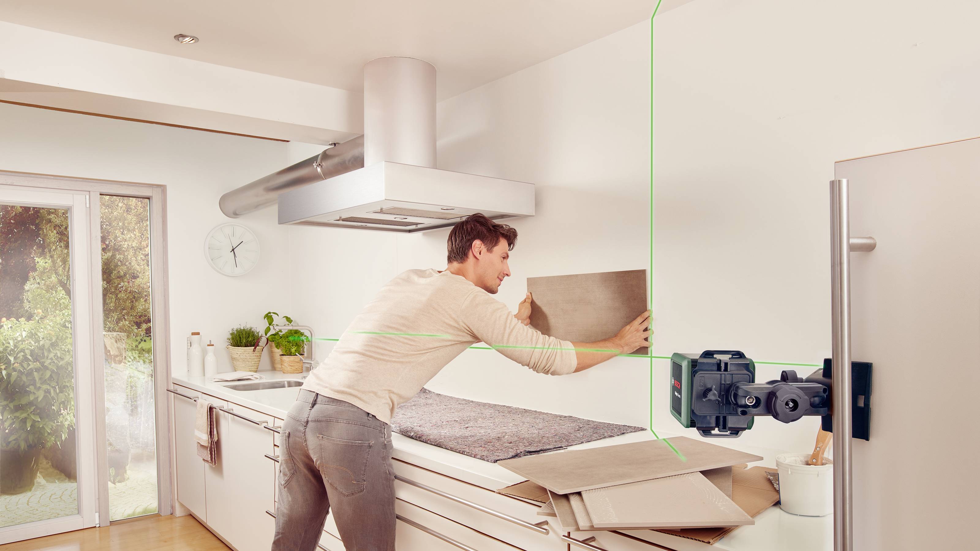 Ein Mann richtet mit einer Laser-Wasserwaage eine Fliese an einer Küchenwand aus. Im Hintergrund eine moderne Küche mit Tageslicht.
