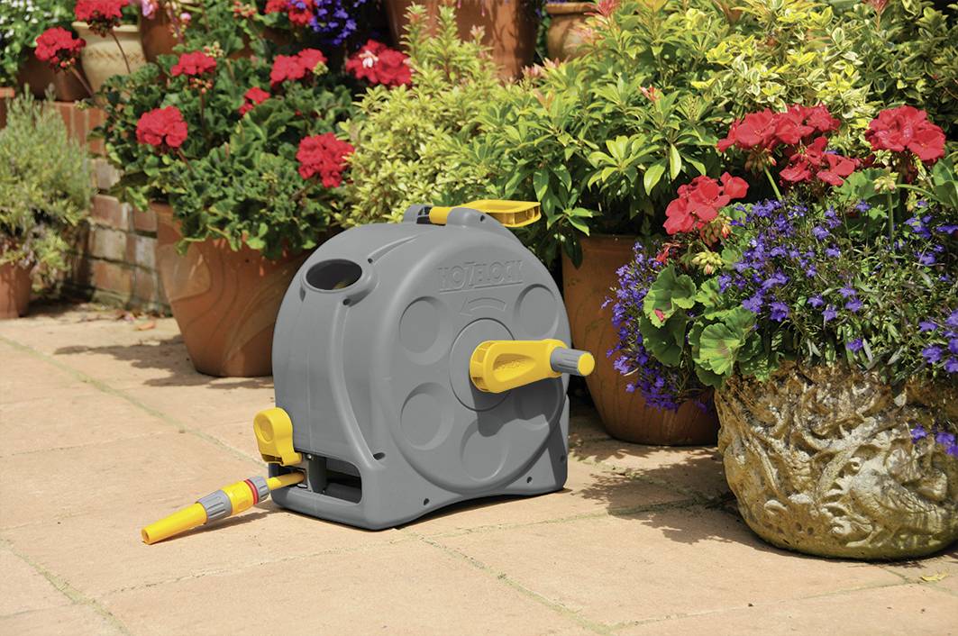 Eine graue Gartenschlauchbox mit gelben Griffen steht auf einer Terrasse, umgeben von blühenden Pflanzen in verschiedenen Töpfen.
