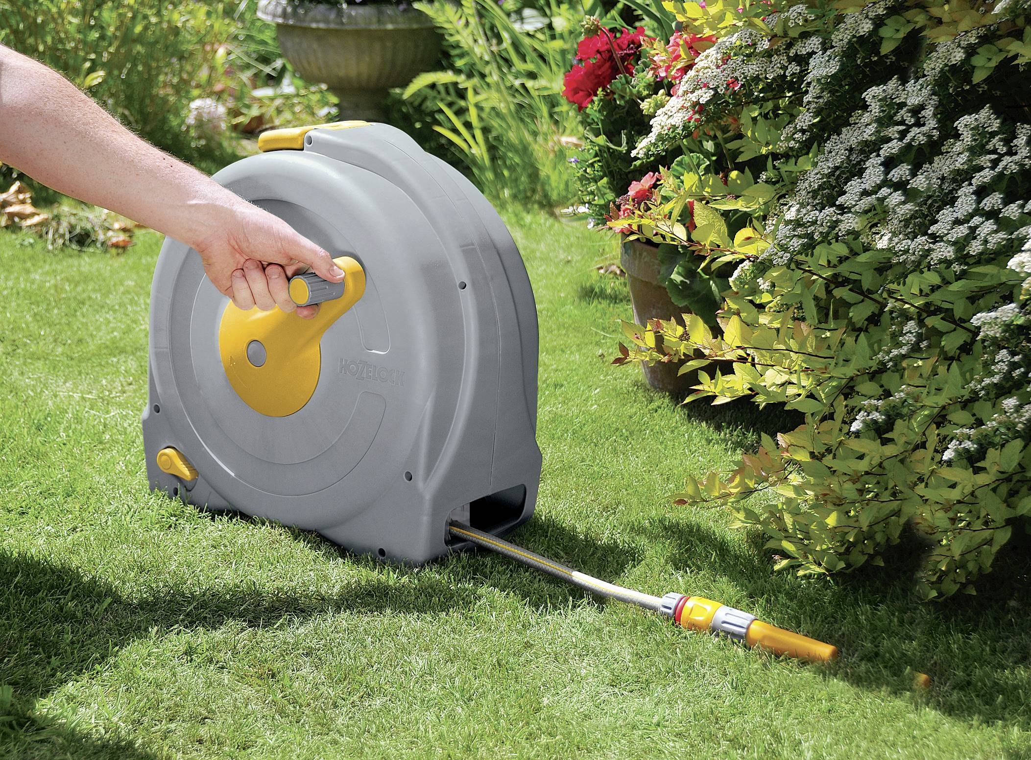Eine Person bedient einen Gartenschlauchaufroller auf einer grünen Wiese, umgeben von Blumen und Sträuchern.