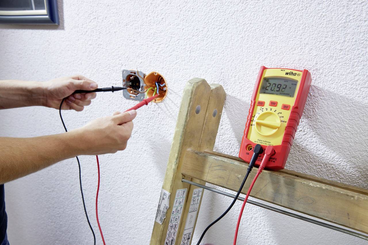 Eine Person misst die Spannung an einer Steckdose mit einem digitalen Multimeter, das auf einer Holzleiter liegt.