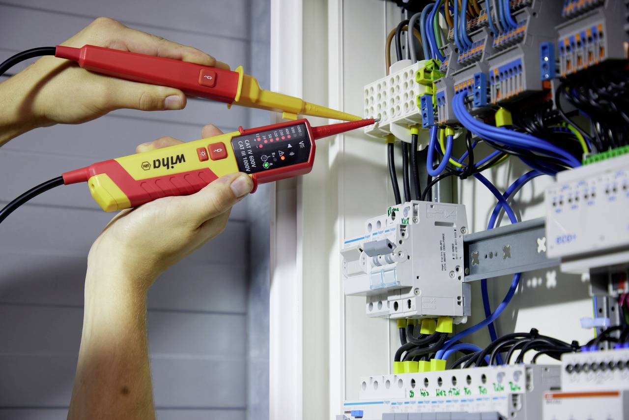 Eine Person misst mit einem elektrischen Prüfgerät die Spannung in einem Schaltschrank mit zahlreichen Kabeln und Schaltungen.