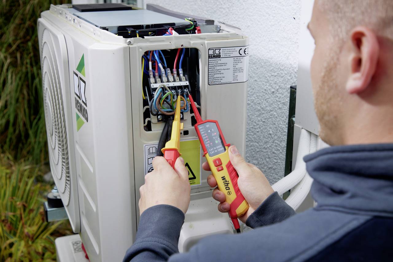 Eine Person überprüft mit einem Multimeter die elektrischen Anschlüsse einer Klimaanlage im Freien.