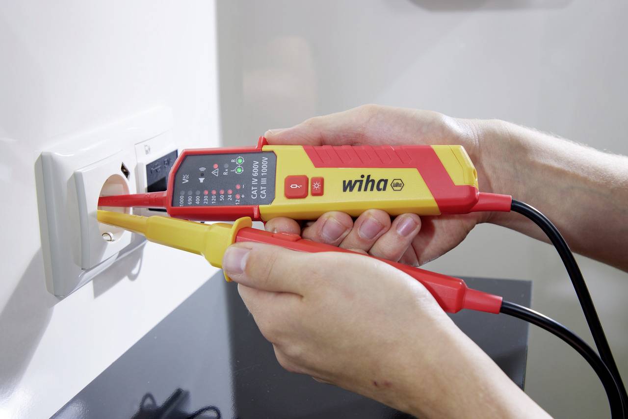 Eine Person misst mit einem Spannungsprüfer die Spannung einer Steckdose. Der Prüfer zeigt ein Messergebnis auf dem Display.