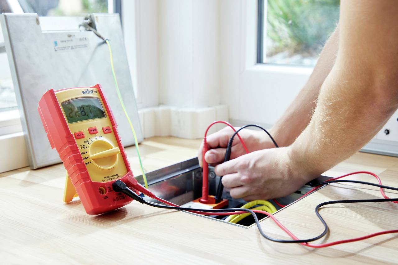 Eine Person misst mit einem digitalen Multimeter die elektrische Spannung in einem Bodenschacht. Kabel sind angeschlossen.