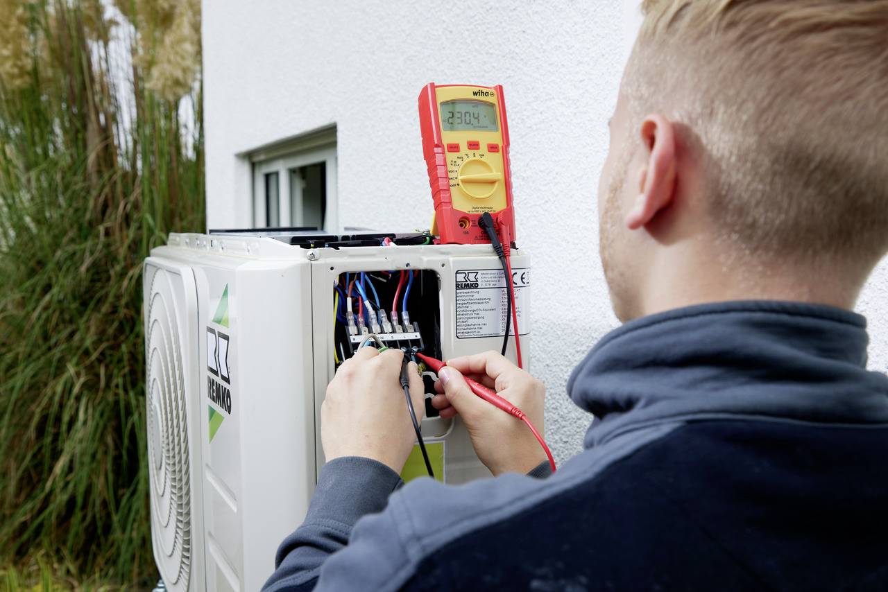 Ein Techniker prüft mit einem Multimeter die Kabelverbindungen einer installierten Wärmepumpe an einer Außenwand.