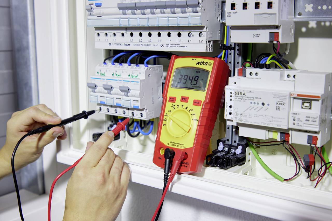 Zwei Hände verwenden ein Multimeter an einem geöffneten Schaltschrank, um elektrische Verbindungen zu testen, mit Fokus auf Messwert 394,9.