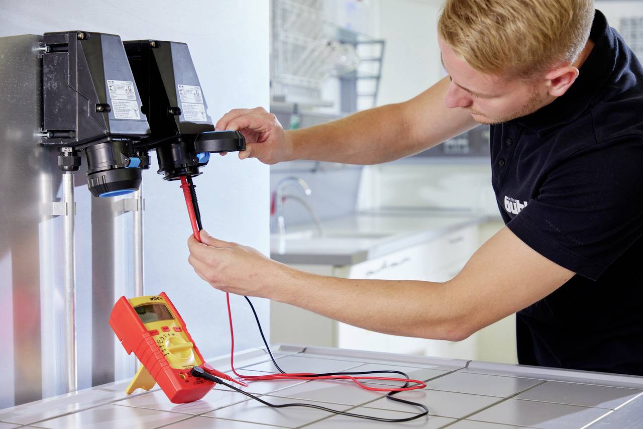 Ein Mann misst elektrische Werte mit einem Multimeter an einer industriellen Maschine in einer Werkstattumgebung.