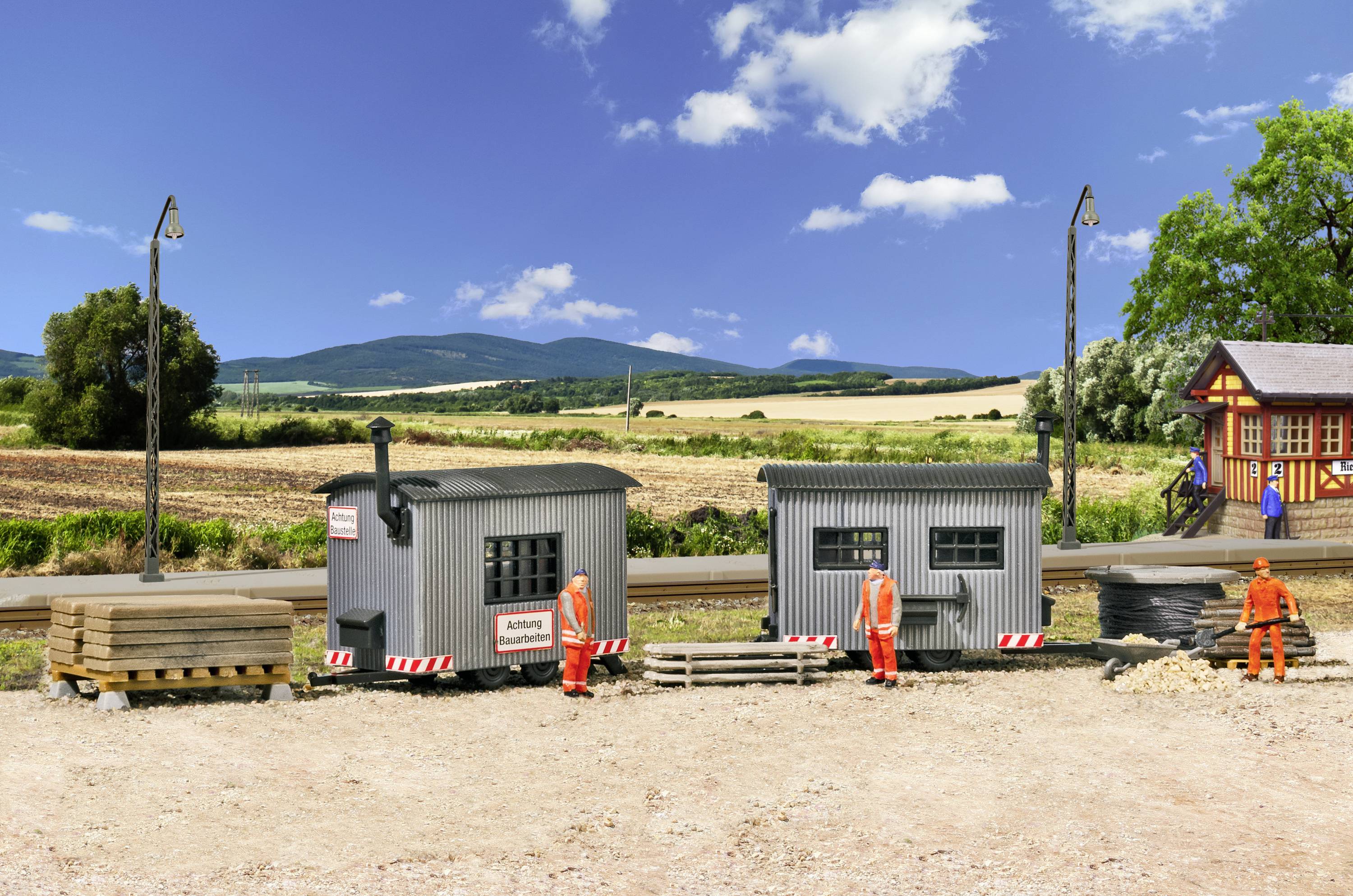 Bauarbeiter in orangefarbener Kleidung arbeiten an einem Baustellenbereich auf einer ländlichen Straße. Hintergrund zeigt Felder und Hügel.