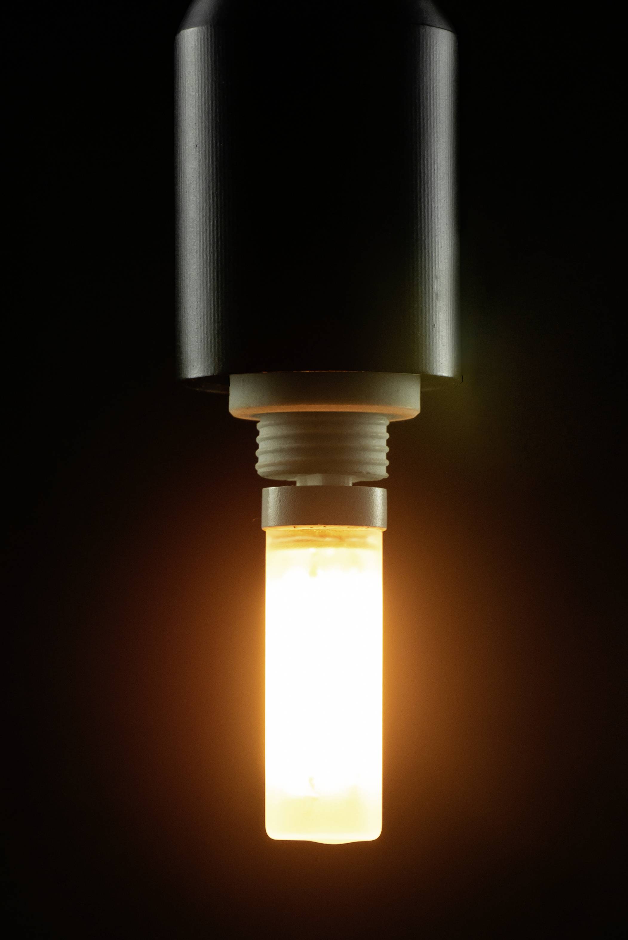 Eine leuchtende Glühlampe hängt von einer schwarzen Fassung, vor einem dunklen Hintergrund. Die Lampe strahlt warmes, orangefarbenes Licht aus.