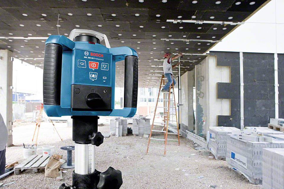 Ein Bauarbeiter arbeitet auf einer Leiter an einer Decke, während im Vordergrund ein blauer Laser-Nivelliergerät steht.