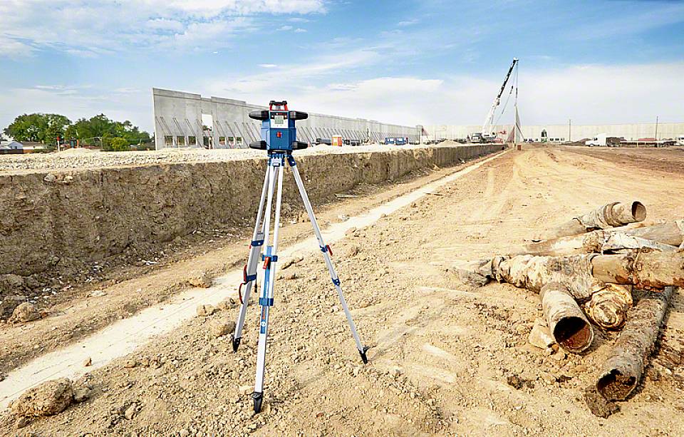Baustelle mit Vermessungsgerät im Vordergrund und Bauarbeiten im Hintergrund, einschließlich Baukränen und Rohren.