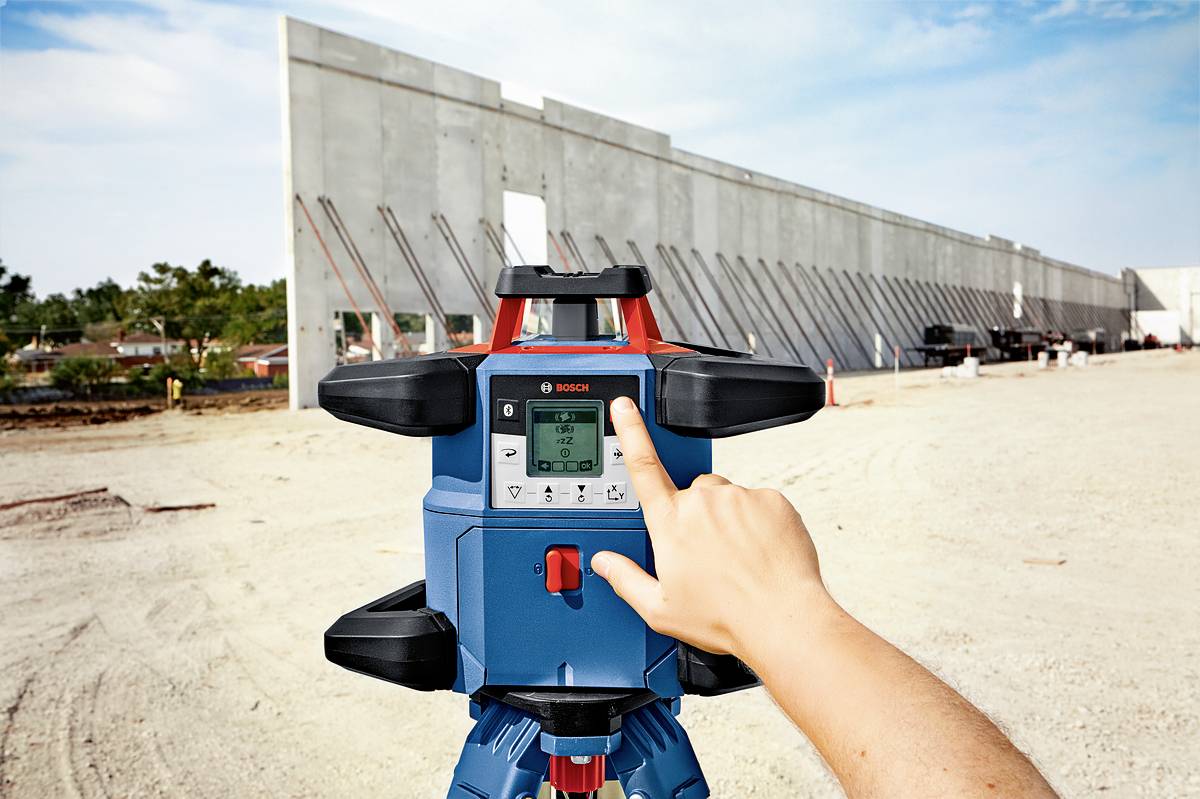 Eine Hand bedient ein Laser-Nivelliergerät auf einer Baustelle vor einer großen Betonwandkonstruktion mit stützenden Streben.
