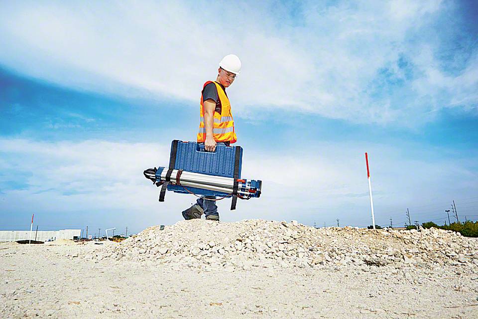 Eine Person in Bauarbeiterkleidung trägt eine große Ausrüstungskiste über eine trockene Baustelle. Der Himmel ist teilweise bewölkt.