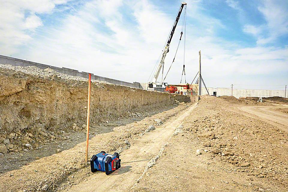Eine Baustelle mit einem Kran, der eine große Betonplatte hebt. Im Vordergrund liegt ein Messgerät auf dem Boden. Im Hintergrund ist ein teilweise errichtetes Bauwerk zu sehen.
