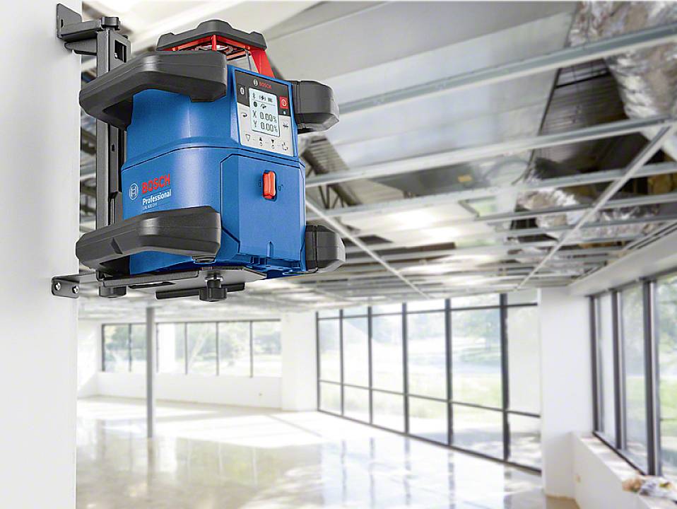 Ein blauer Laser-Nivelliergerät ist an einer Wandhalterung in einem modernen Büro mit großen Fenstern und sichtbaren Deckeninstallationen befestigt.