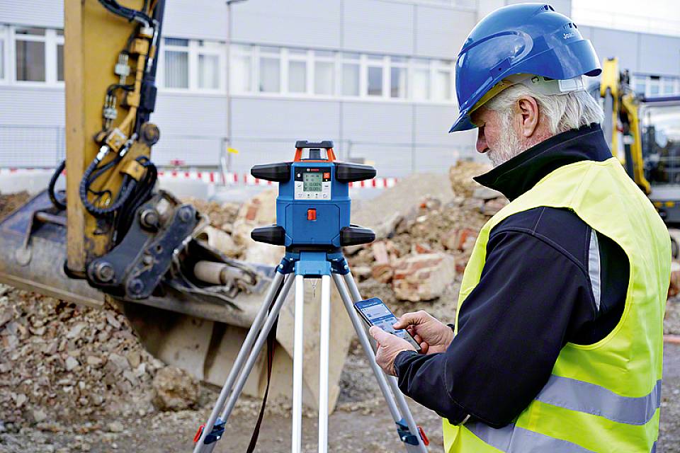 Ein Bauarbeiter mit Schutzhelm und Warnweste bedient ein Vermessungsgerät auf einer Baustelle, im Hintergrund ist ein Bagger.