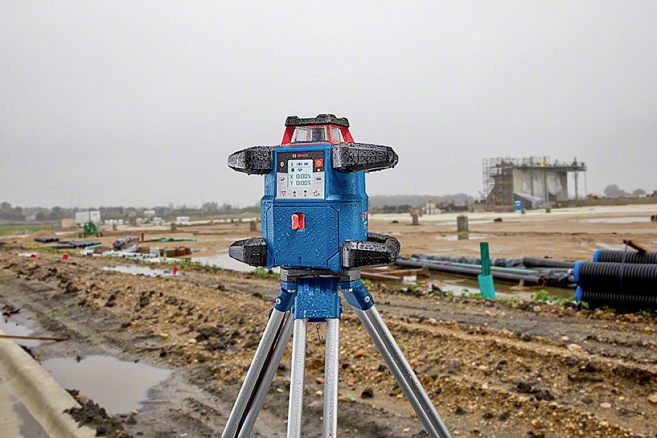 Ein blaues Vermessungsgerät auf einem Baustativ im Vordergrund auf einer Baustelle; unscharfe Hintergrund zeigt Rohre und Baugerüste.
